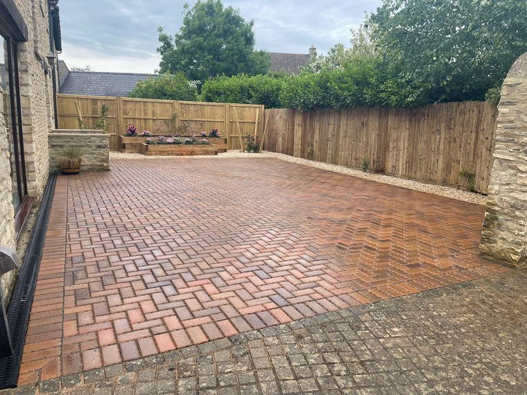 A wide block-paved courtyard enclosed by close-board fencing and a Cotswold stone wall