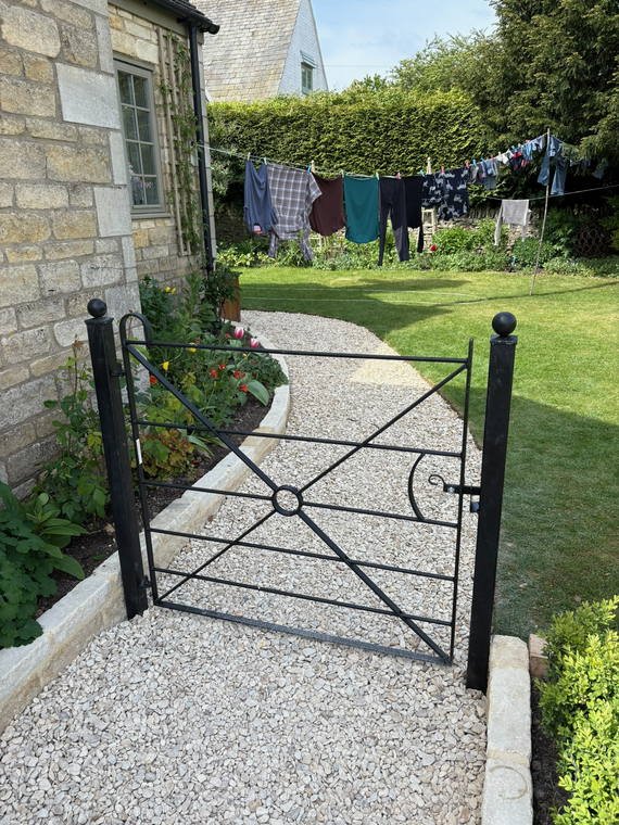 An ornate metal gate opening onto a gravel path beside a Cotswold cottage