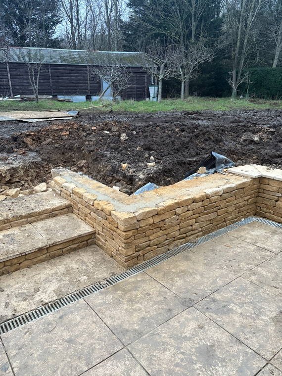 A Cotswold stone retaining wall under construction beside porcelain paving and a linear drain